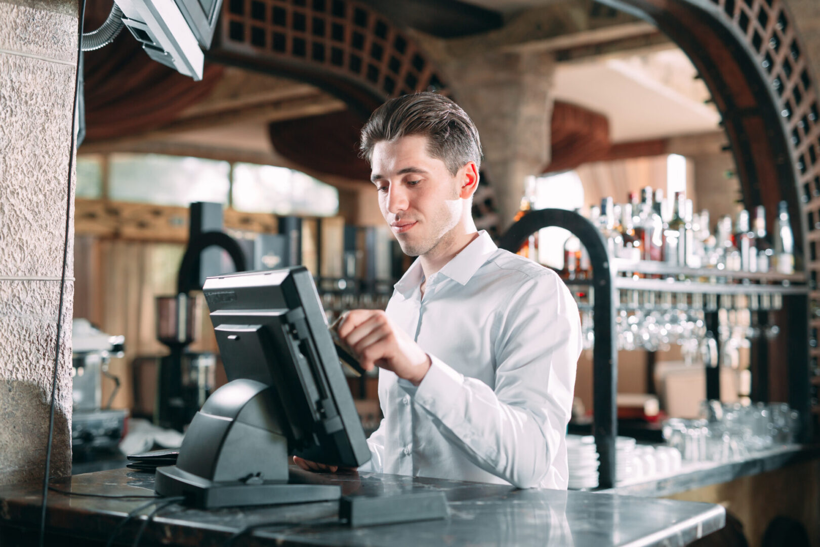 small business, people and service concept - happy man or waiter in apron at counter with cashbox working at bar or coffee shop